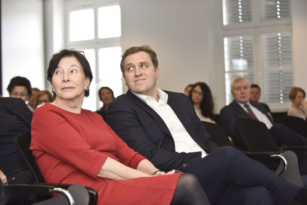 Eva Luise Köhler und ein Mann neben ihr schauen sitzend nach hinten links, im Hintergrund sieht man verschwommen das Publikum