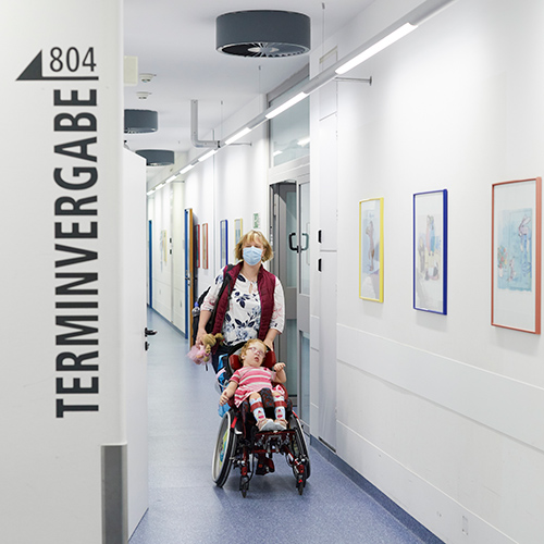 Eine Frau schiebt einen Rollstuhl mit einem offensichtlich schwerbehinderten kleinen Mädchen durch einen Krankenhausgang. In derHand hält sie eine Puppe. Sie nähern sich einem Zimmer, das mit 