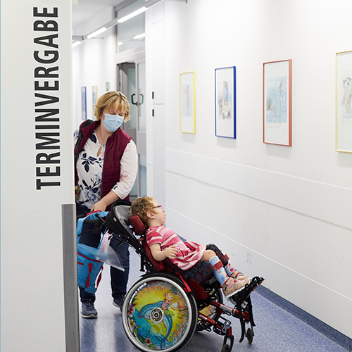 Eine Frau Schiebt ein Kind im Rollstuhl durch den Flur eines Krankenhauses.