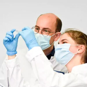 Ein Mann und eine Frau, beide mit Mund-Nasen-Bedeckung, Kittel und blauen Einweg-handschuhen, stehen dicht beieinander und prüfen einen Objektträger.
